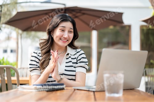 Preview: Happy asian woman clapping hands as looking at a laptop while sitting at wooden table in garden cafe
