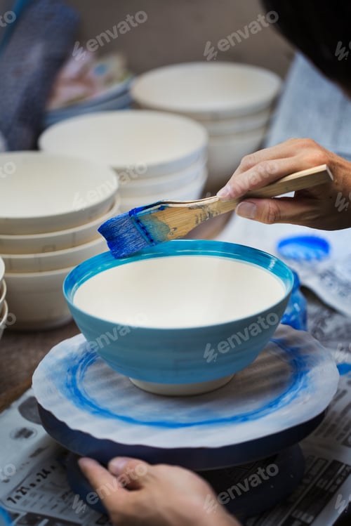 Preview: Close up of person working in a Japanese porcelain workshop, painting white bowls with blue glaze.