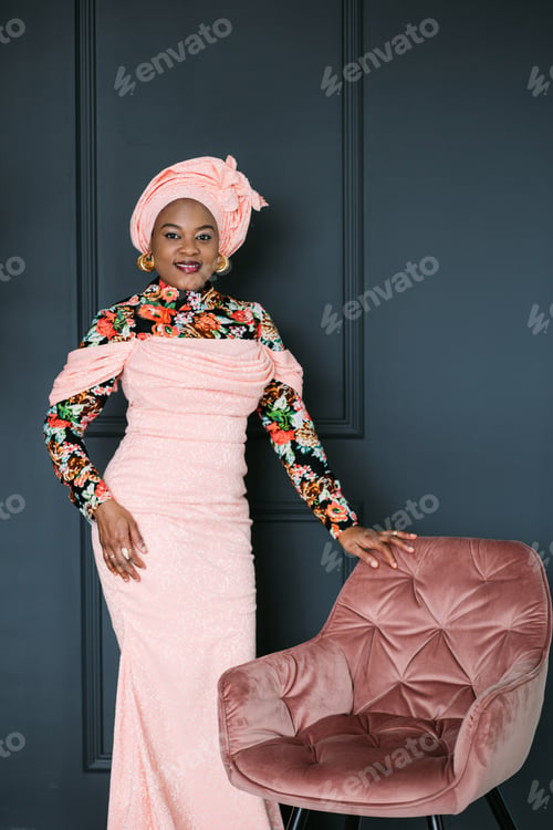 Preview: Gorgeous african woman in traditional dress and headscarf, standing in studio near pink soft chair