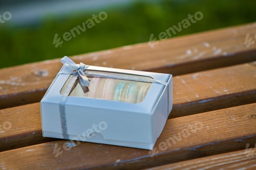 Preview: Cardboard white gift box with colorful handmade macaron cookies on wooden bench outdoors