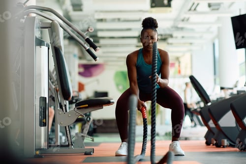 Preview: Black female athlete exercising with battle ropes while working out in a gym.