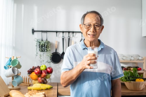 Preview: Asian senior mature man drinking a glass of milk in kitchen at home.