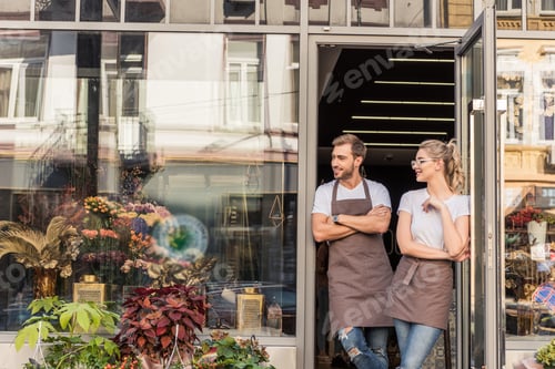 Preview: cheerful caucasian florists standing near flower shop and looking away