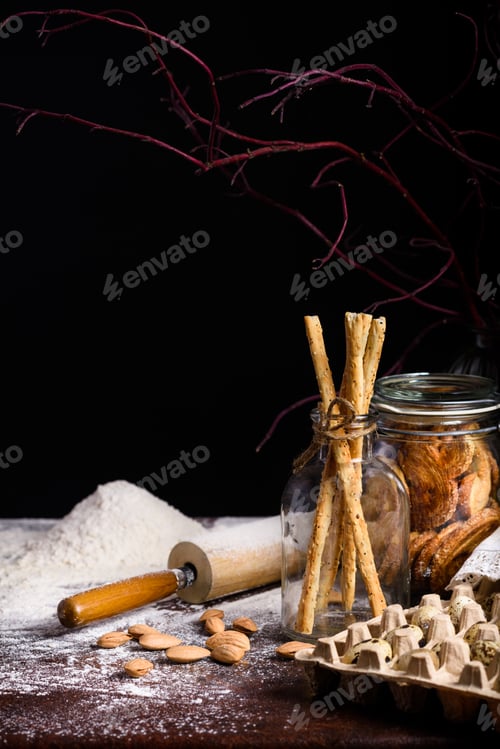 Preview: close-up view of egg box, cookies, breadsticks and rolling pin with flour on table top on black