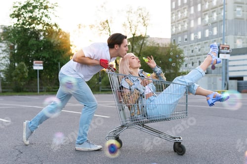 Preview: Couple have fun with a shopping trolley and blowing bubbles on a supermarket parking