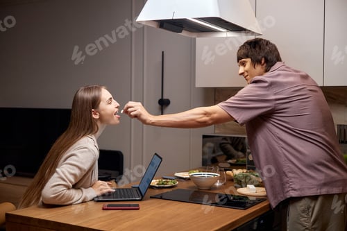 Preview: Young Man Feeding Young Woman Working on Laptop in Kitchen
