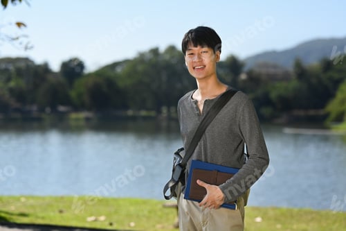 Preview: Portrait of asian student man standing near lake with mountains on background.