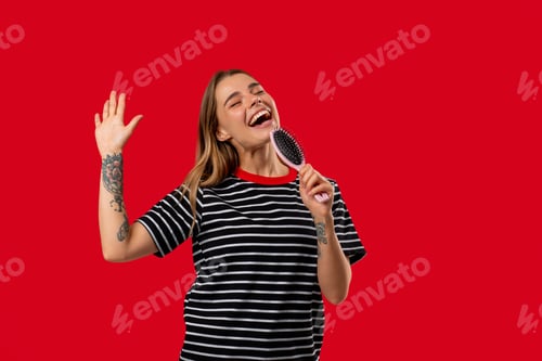 Preview: Playful Young Woman Singing and Dancing With Hairbrush On Red Studio Background