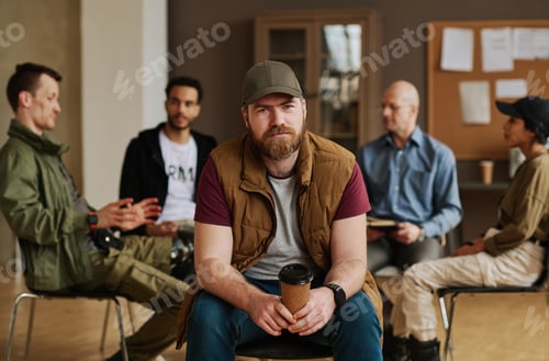 Preview: Bearded man with cup of coffee looking at camera against group of people
