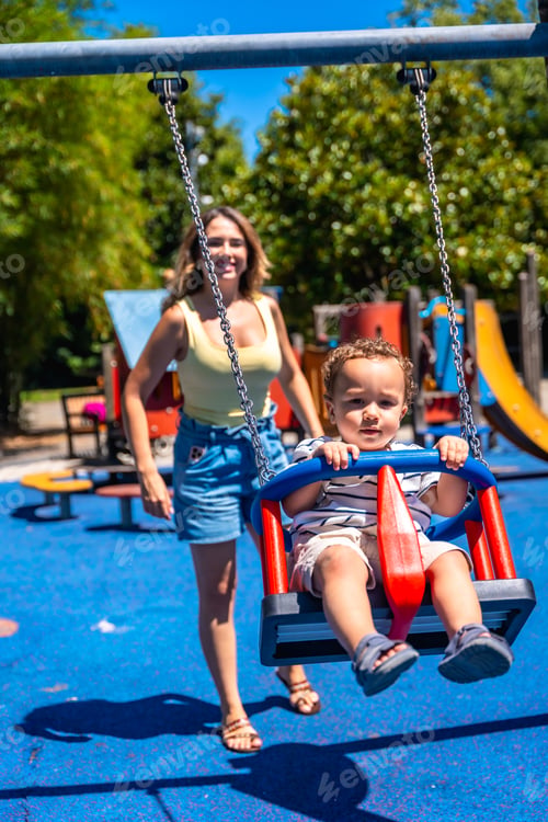 Preview: Happy toddler swinging at playground with mother watching