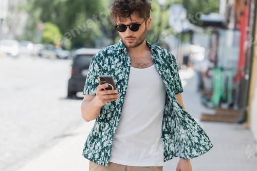 Brunette man in sunglasses using smartphone while walking on urban street