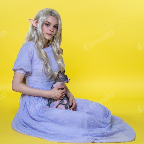 Preview: Young blonde woman cosplay elf sitting on yellow background, holding Sphinx kitten on her lap