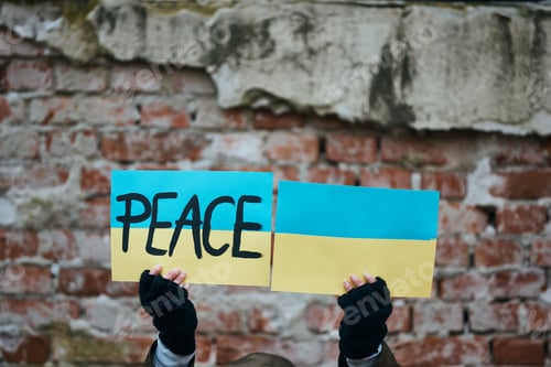 Preview: Close-up of person holding Ukrainian flags with peace message against war damaged wall.