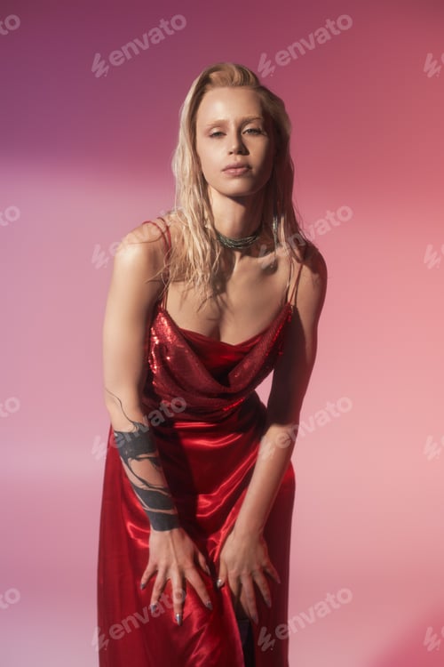 Preview: beautiful stylish woman in fashionable red dress posing on pink backdrop and looking at camera