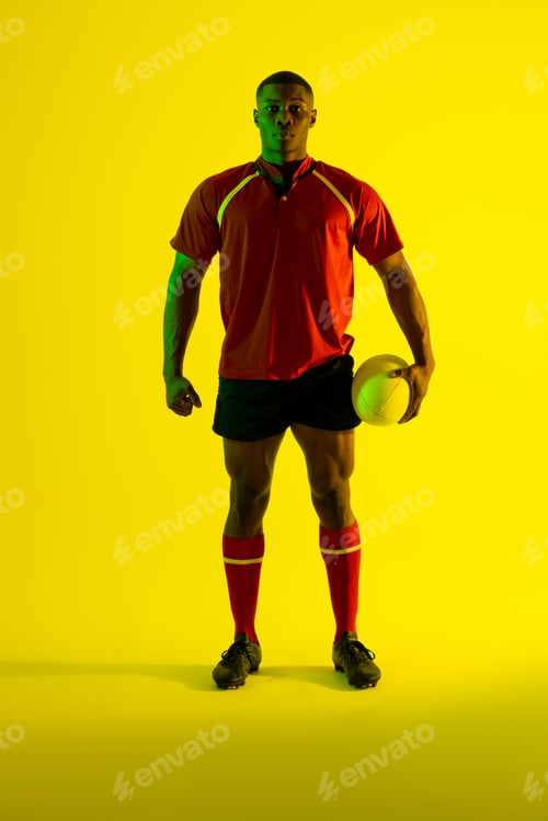 Preview: Portrait of african american male rugby player with rugby ball over yellow lighting