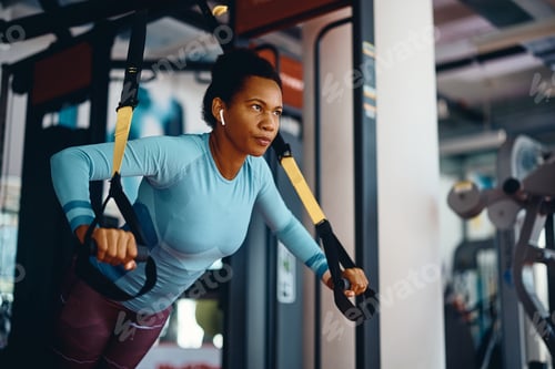 Preview: Black sportswoman doing TRX exercises with suspension straps in a gym.