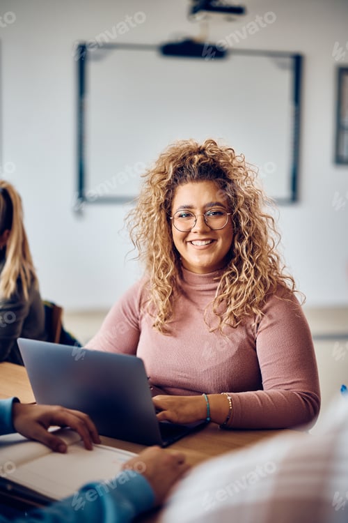 Preview: Young happy woman studying on laptop at university classroom and looking at camera.