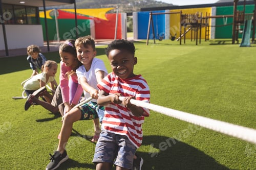 Preview: Front view of multi ethnic group of happy school kids playing tug of war in playground