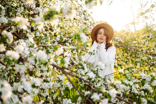 Preview: Portrait of Woman In hat posing near flowering tree. Smiling young woman enjoying smell of flowers.