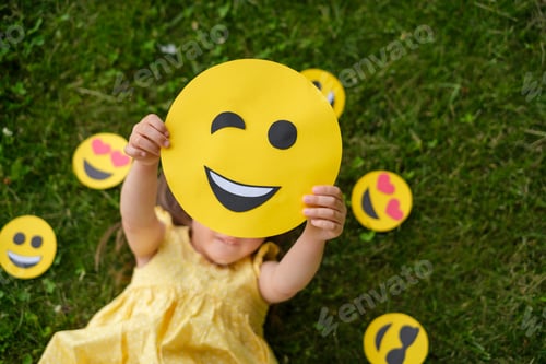 Preview: Girl holds paper cute winking emoticon lying on grass of lawn