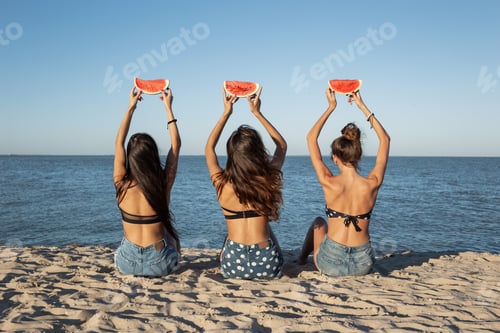 Preview: Back view of three dark-haired girls in jean shorts sitting on the sand near the sea holding