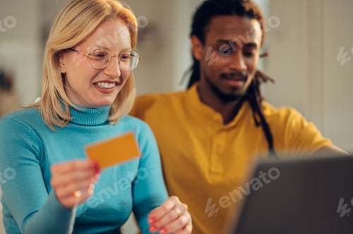 Preview: A happy blonde is using her credit card for online booking on the laptop while sitting with husband.