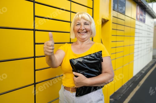 Preview: Senior 50s woman showing thumb up getting parcel from cell of automatic post terminal outdoors