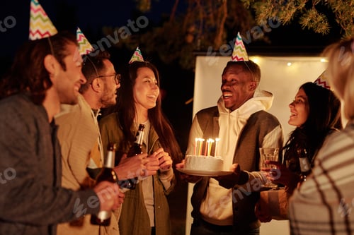 Preview: Young African man with birthday cake looking at his friends toasting with drinks