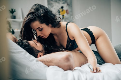 Preview: passionate woman in black underwear holding condom in hands near boyfriend lying on bed