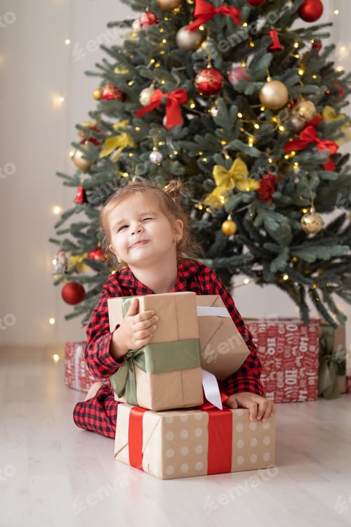 Preview: cute little child girl in red pajama with gift boxes sitting under christmas tree at home