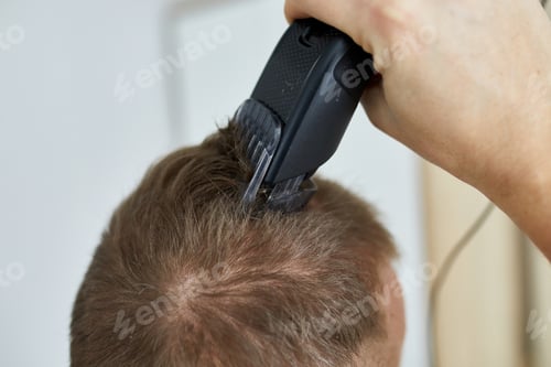 Preview: Handsome man in white t-shirts cutting hair personally himself with machine trimmer, clipper at home