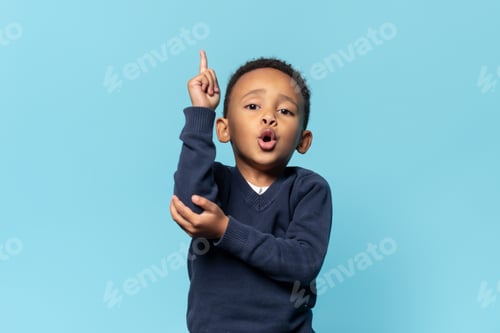 Preview: Funny black kid little boy holding finger up and looking at camera, showing new idea on blue studio