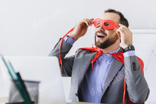Preview: excited super businessman in cape looking through red mask in office