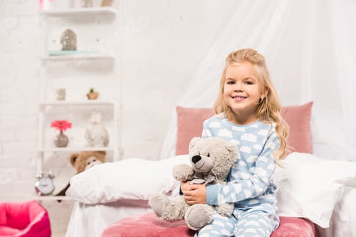 Preview: smiling cute kid sitting on bed with teddy bear and looking at camera