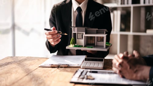 Preview: A man is holding a model house and a pen on a table