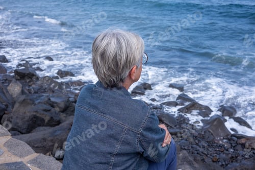 Preview: Rear view of white-haired senior woman sitting on the beach wearing eyeglasses and denim jacket