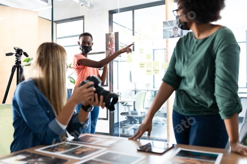 Preview: Diverse group of business colleagues wearing masks brainstorming holding camera in meeting room