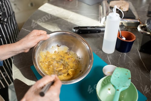 Preview: Hands of mature female mixing raw ingredients for stew with whisker in metallic bowl while cooking
