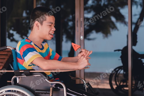 Preview: Smart disabled child on wheelchair eating watermelon on summer sea beach.