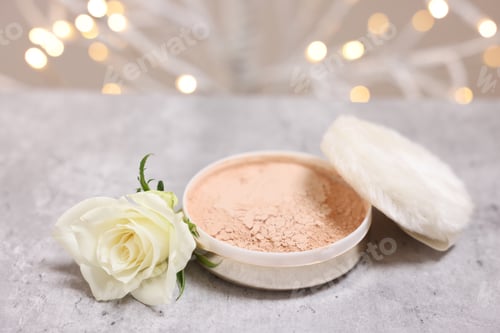 Preview: Face powder, puff applicator and rose flower on grey textured table against blurred lights, closeup