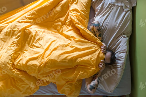 Preview: Tattooed man lying in bed, blanket over his face