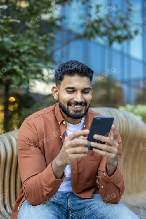 Preview: Young man smiling using smartphone sitting outdoors