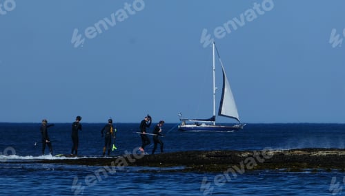 Preview: group of young men in wetsuits carrying spearfishing gear with sailboat on water against blue sky