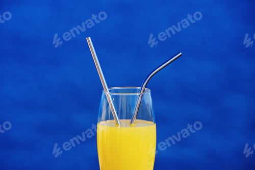 Preview: Closeup view of drinking glass with orange juice and metallic straws against blue sea background