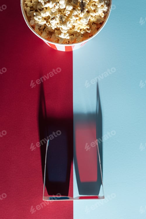 Preview: top view of cardboard 3d glasses with long vertical shadow and striped bucket with popcorn on blue