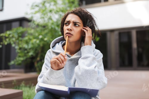 Preview: Pensive lady with dark curly hair sitting with book on knees and pencil in hand
