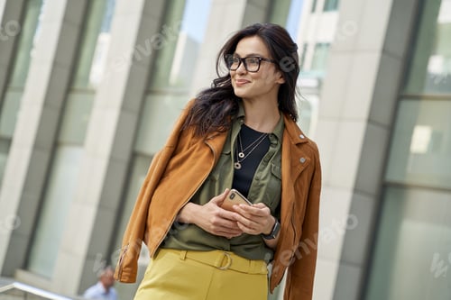 Preview: Young stylish woman holding smartphone, looking aside and smiling while standing against blurred