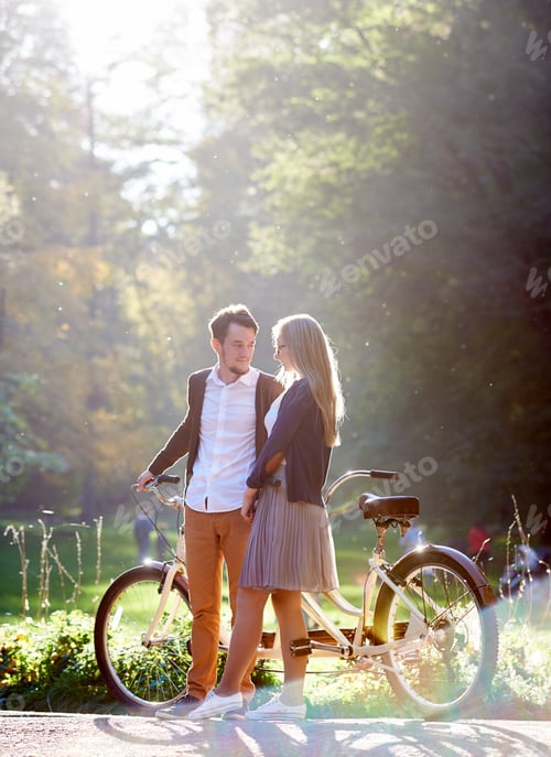 Preview: Young couple, handsome man and attractive woman on tandem bike in sunny summer park or forest.