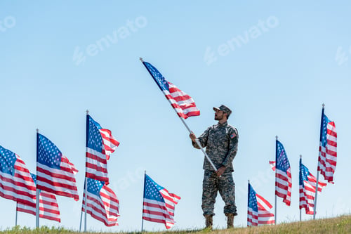 Preview: selective focus of handsome soldier in military uniform and cap holding american flag