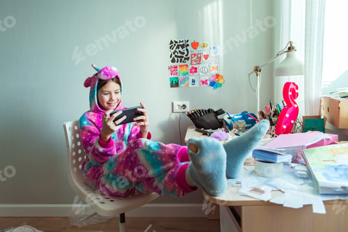 Preview: Child girl using phone, playing phone games in messy childrens room. Kid playing among the many toys